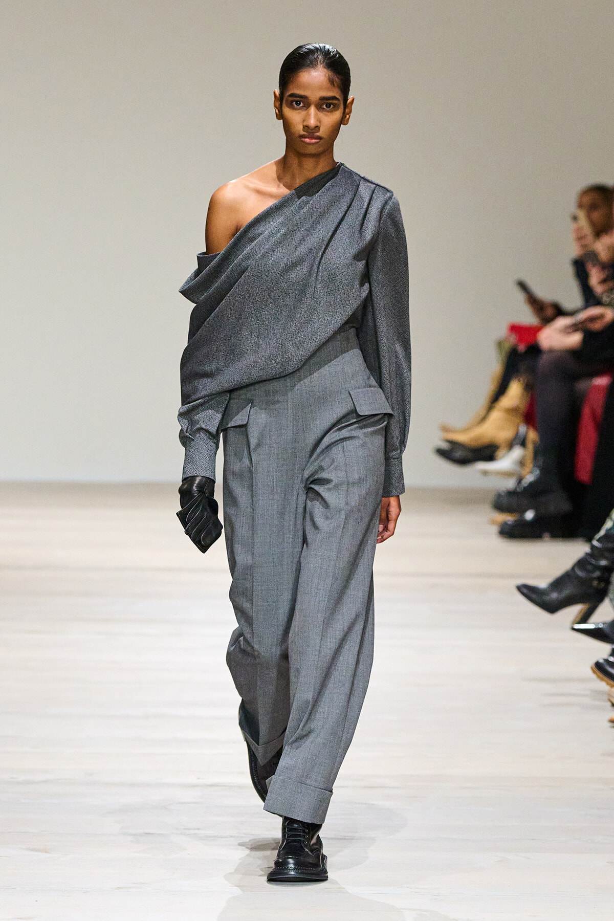 Female model walking a runway in a gray one-shoulder top with matching wide pants, wearing a black glove on her left hand.