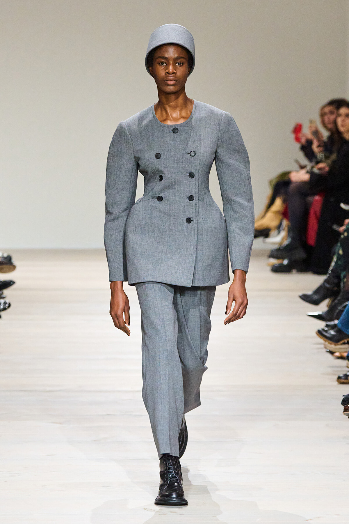 Model walks the runway in a light gray double-breasted suit with structured shoulders, matching straight-leg pants, black lace-up boots, and a gray hat.