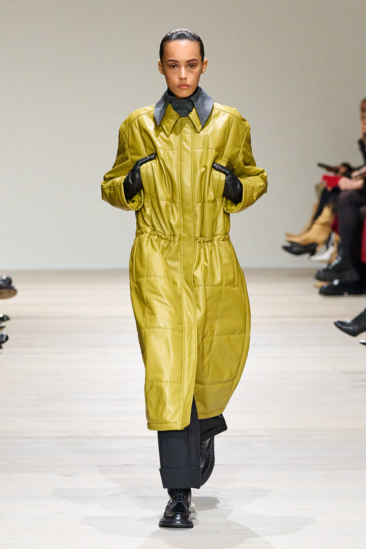 Model walks a runway wearing a mustard yellow quilted coat with black gloves and dark cropped pants.