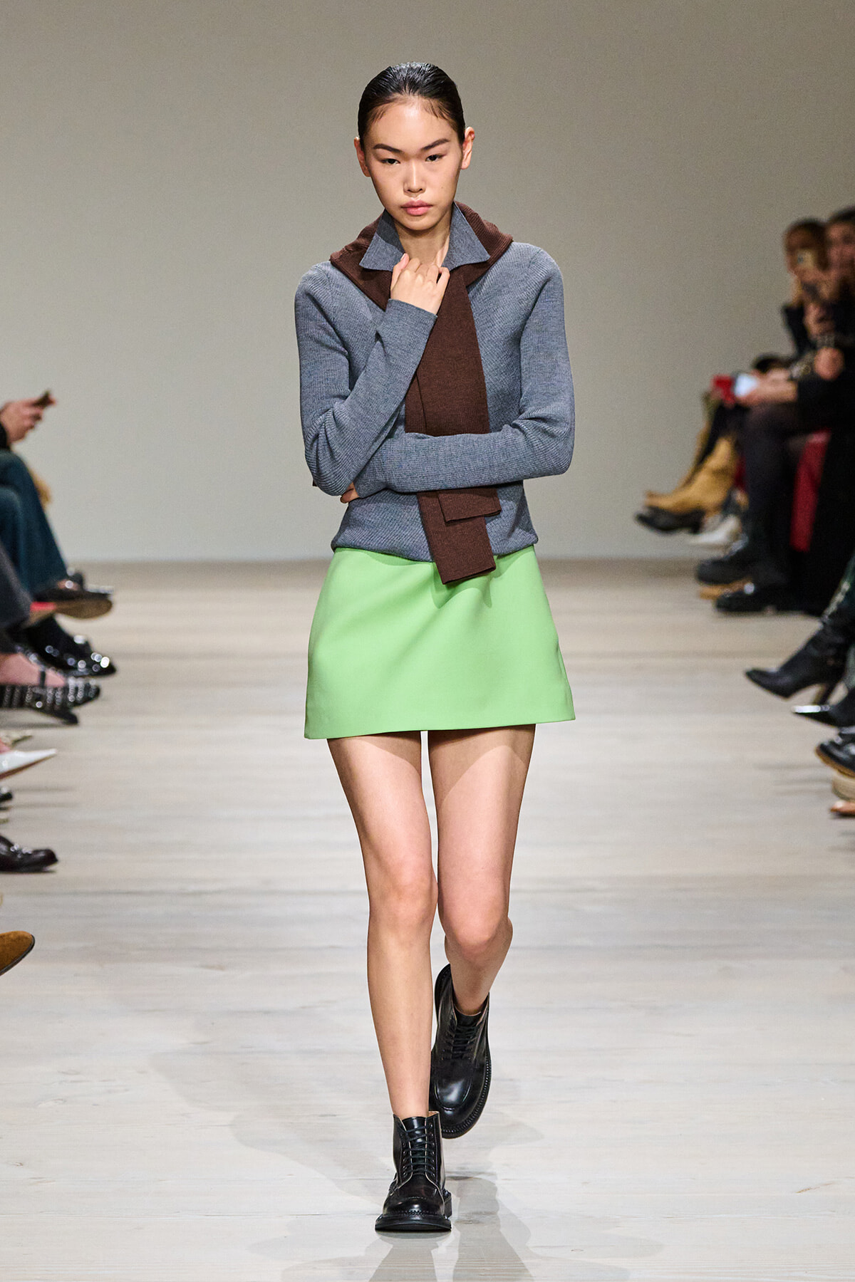 Model on a runway wearing a gray knit sweater, brown scarf, lime green A-line skirt, and black lace-up boots.