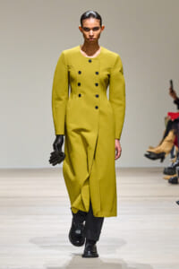 Model walks a fashion runway wearing a mustard-yellow, double-breasted coat with structured shoulders and a high waist, paired with black gloves and boots.