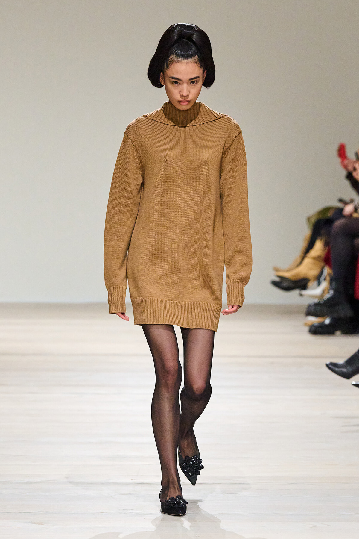 Model walking runway in a camel knit sweater dress, sheer black tights, and black embellished flats; hair styled in a large updo.