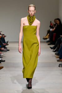 Model walks a runway in a strapless olive-green midi dress with a matching cowl neck scarf accessory and black heels as audience watches.