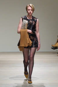 Model walking a runway in a sleeveless dark floral romper, carrying a tan sweater and a newspaper under her arm.