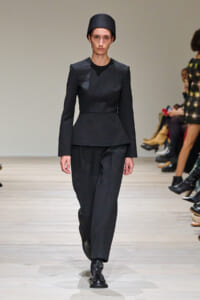Model walks a minimalist black look: tailored peplum jacket, wide-leg trousers, black lace-up boots, and a tall black hat on a runway.