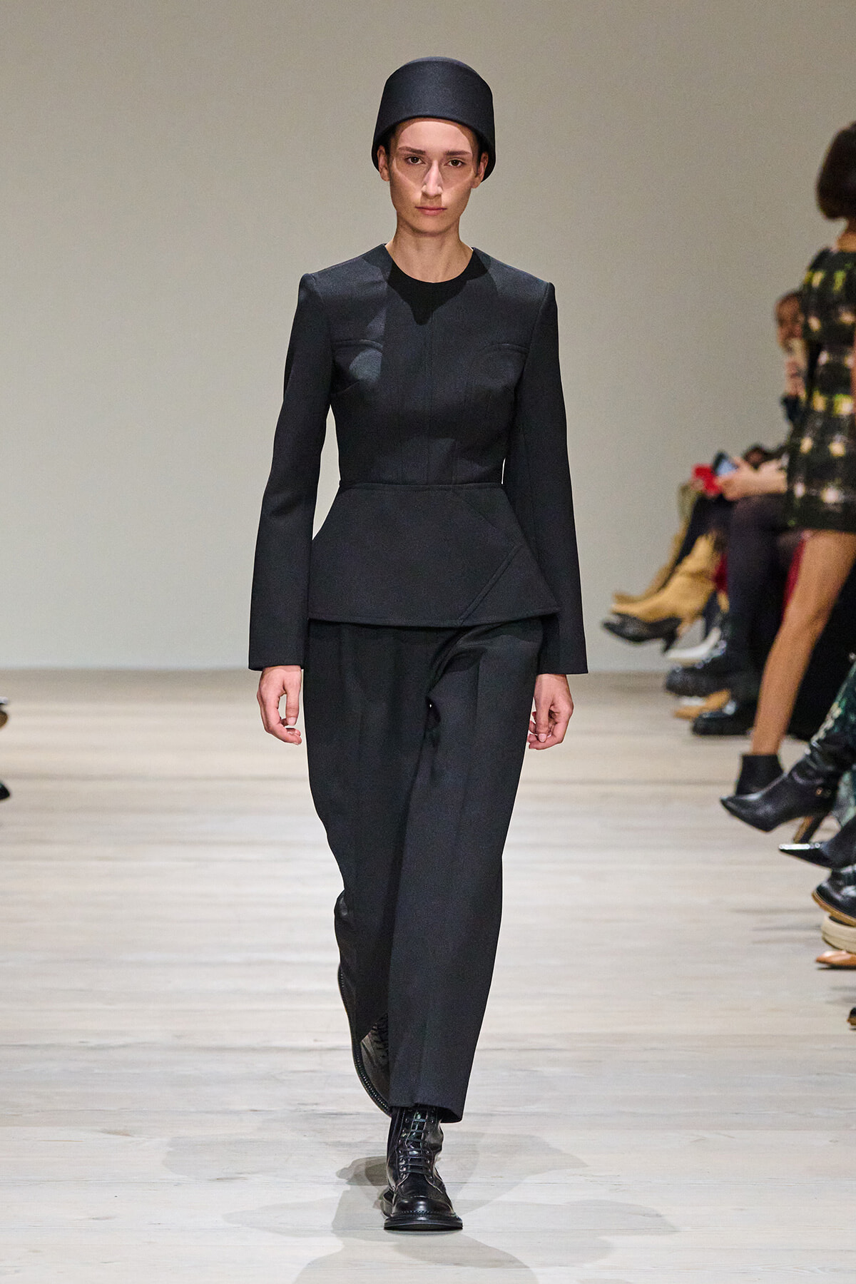 Model walks a minimalist black look: tailored peplum jacket, wide-leg trousers, black lace-up boots, and a tall black hat on a runway.