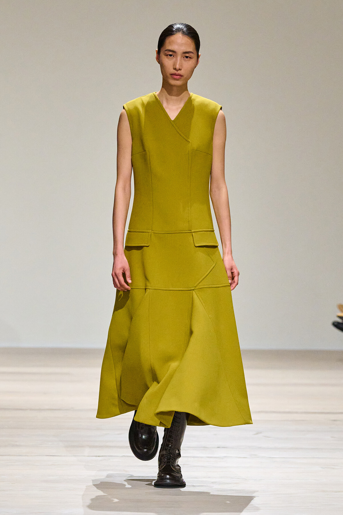Model walking the runway in a mustard sleeveless V-neck structured dress with front pockets and black boots