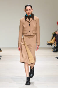 Model walking a beige tailored suit with cropped jacket and asymmetric skirt on a runway, wearing a teal collar under the jacket, black boots.