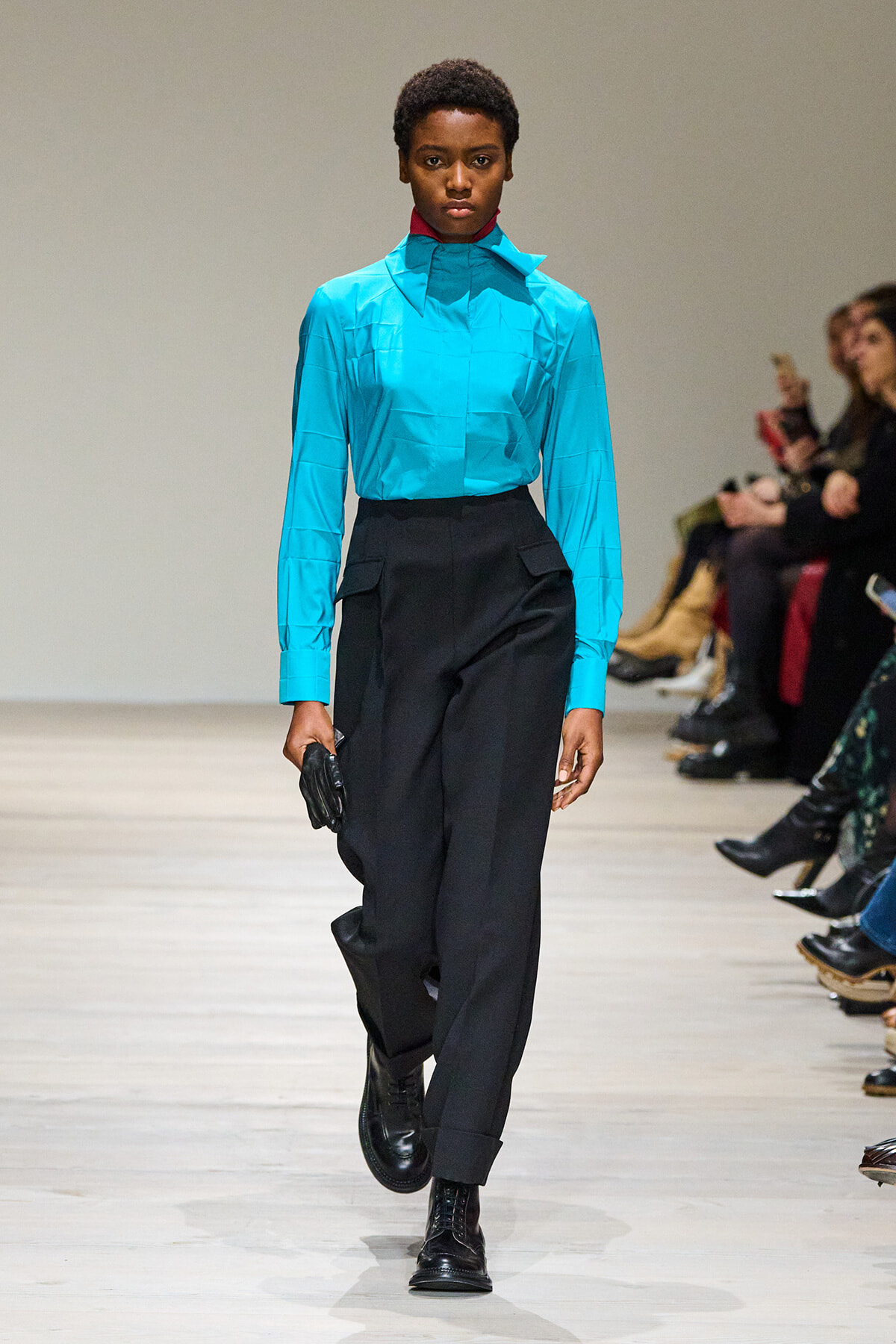 Model walks a runway in a bright turquoise shirt with a high collar and black high-waisted trousers, wearing black boots and carrying a glove in one hand.