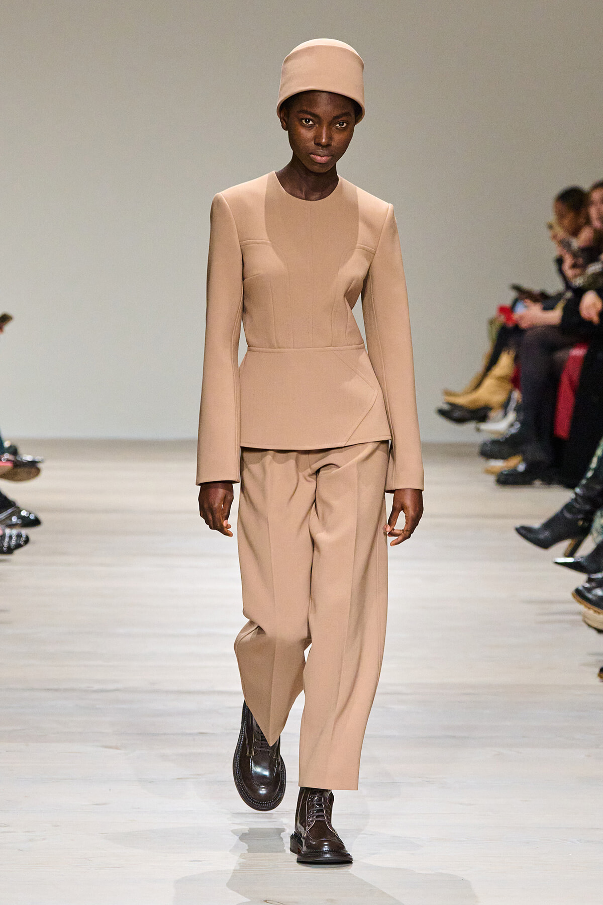 Model on a runway wearing a beige structured top with matching wide-leg pants and a beige hat.