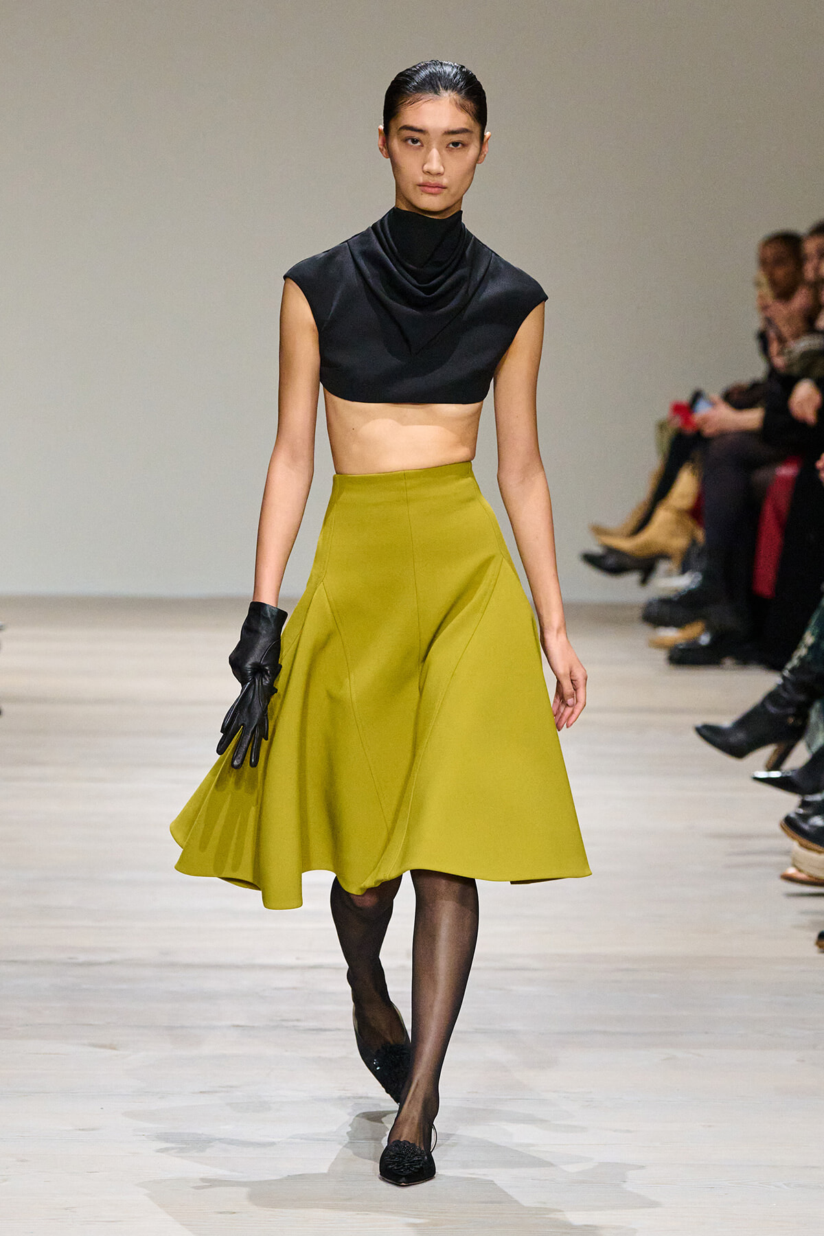 Model on a runway wearing a black draped crop top and an olive midi A-line skirt, with black gloves and sheer tights.