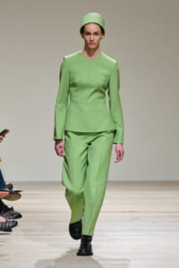 Model walks the runway in a lime-green tailored peplum top and matching trousers, with a matching headwrap and black shoes.