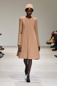 Model wearing a tan A-line coat and matching helmet-style hat on a runway runway show.