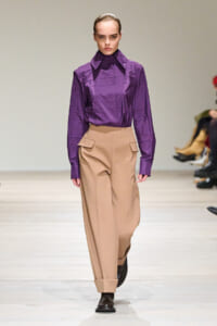 Female model on a runway wearing a purple long-sleeve blouse tucked into beige wide-leg trousers with folded cuffs.