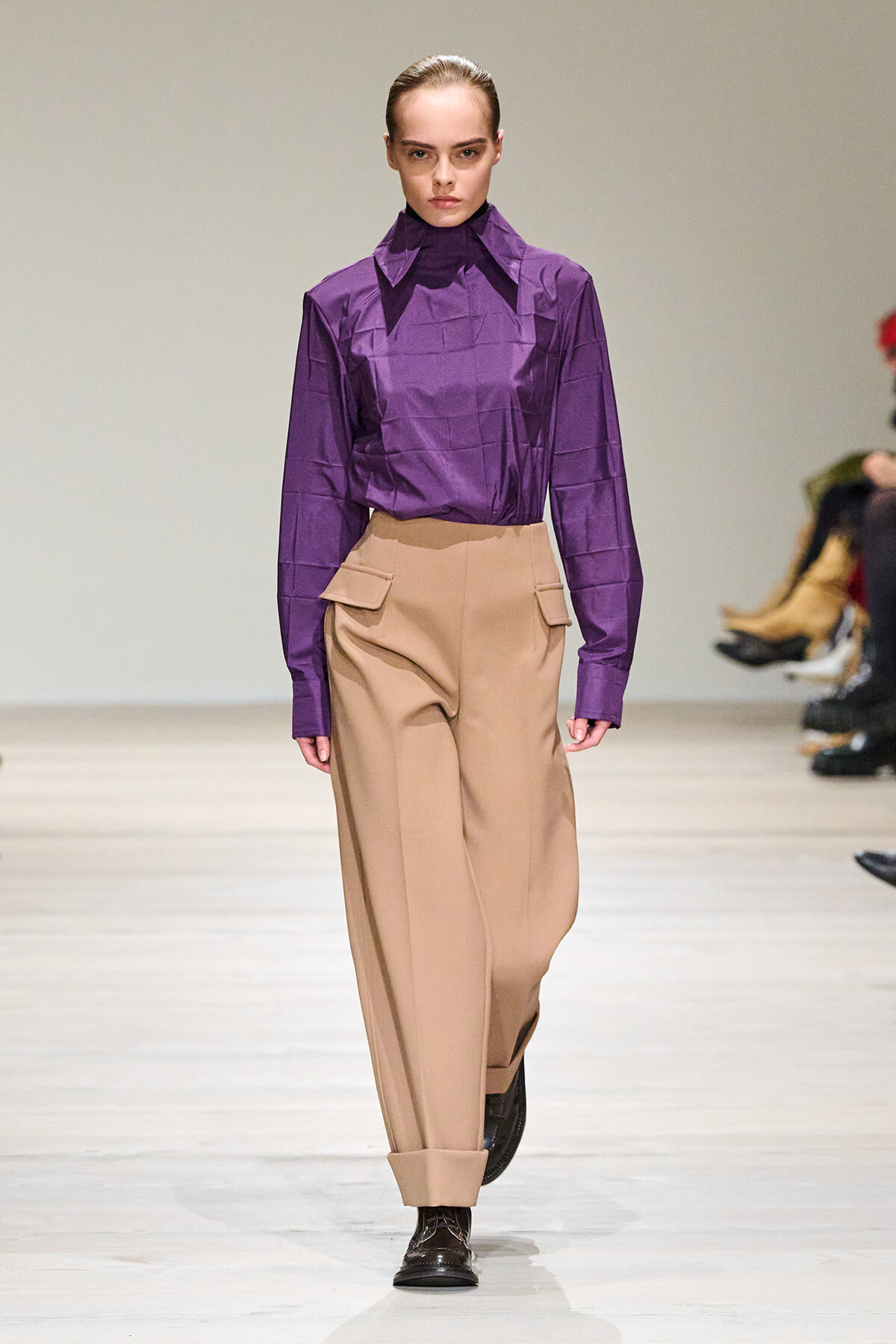 Female model on a runway wearing a purple long-sleeve blouse tucked into beige wide-leg trousers with folded cuffs.