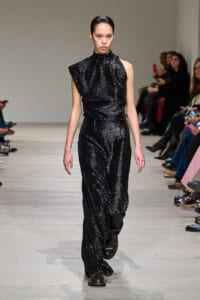 Model walking down a fashion runway in a black sequined jumpsuit with a high ruffled neckline; seated audience along the sides of the catwalk.