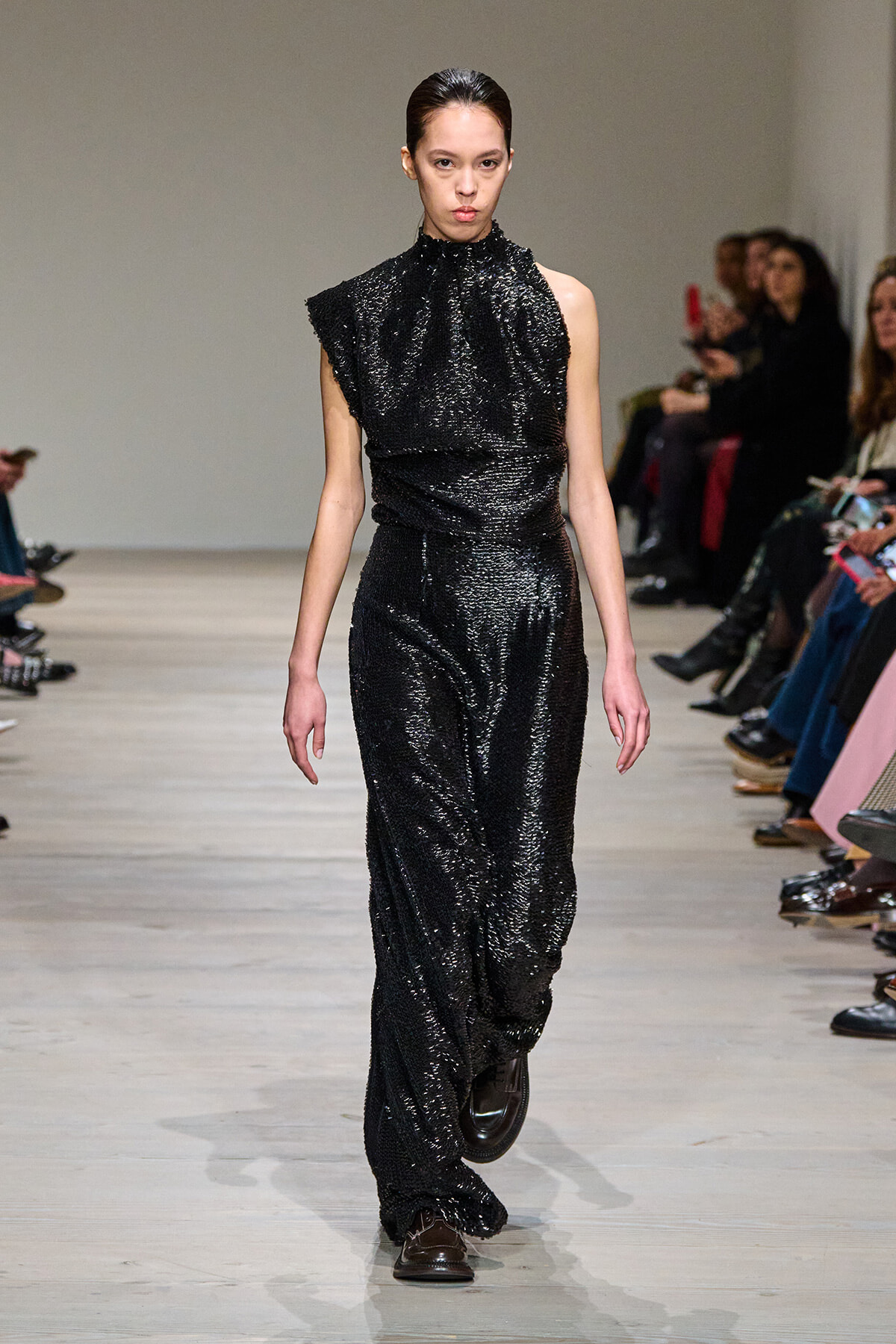 Model walking down a fashion runway in a black sequined jumpsuit with a high ruffled neckline; seated audience along the sides of the catwalk.