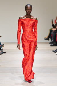 Tall Black model walking the runway in a red off-the-shoulder, floor-length gown with a subtle damask pattern.