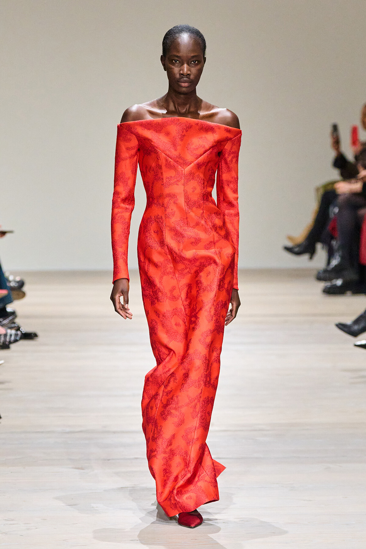 Tall Black model walking the runway in a red off-the-shoulder, floor-length gown with a subtle damask pattern.