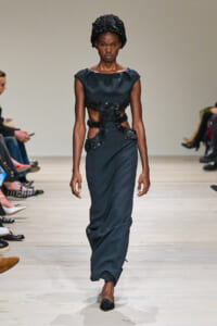 Model on a fashion runway wearing a dark gown with floral appliqué panels at the waist and midriff, plus a textured black headwrap.