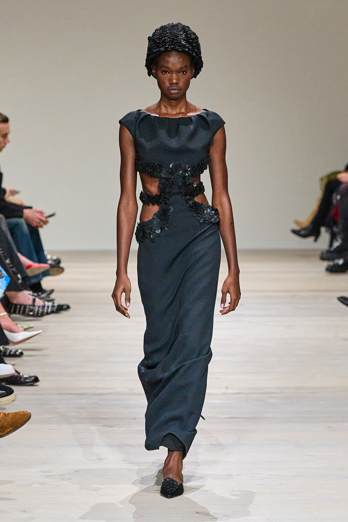 Model on a fashion runway wearing a dark gown with floral appliqué panels at the waist and midriff, plus a textured black headwrap.