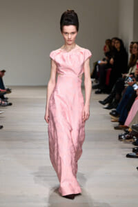 Model walks the runway in a pale pink, off-the-shoulder gown with a textured jacquard pattern and a long, flowing skirt.