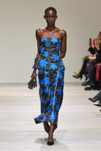 Model on the runway wearing a strapless black top with a blue floral print and matching skirt, holding black gloves.