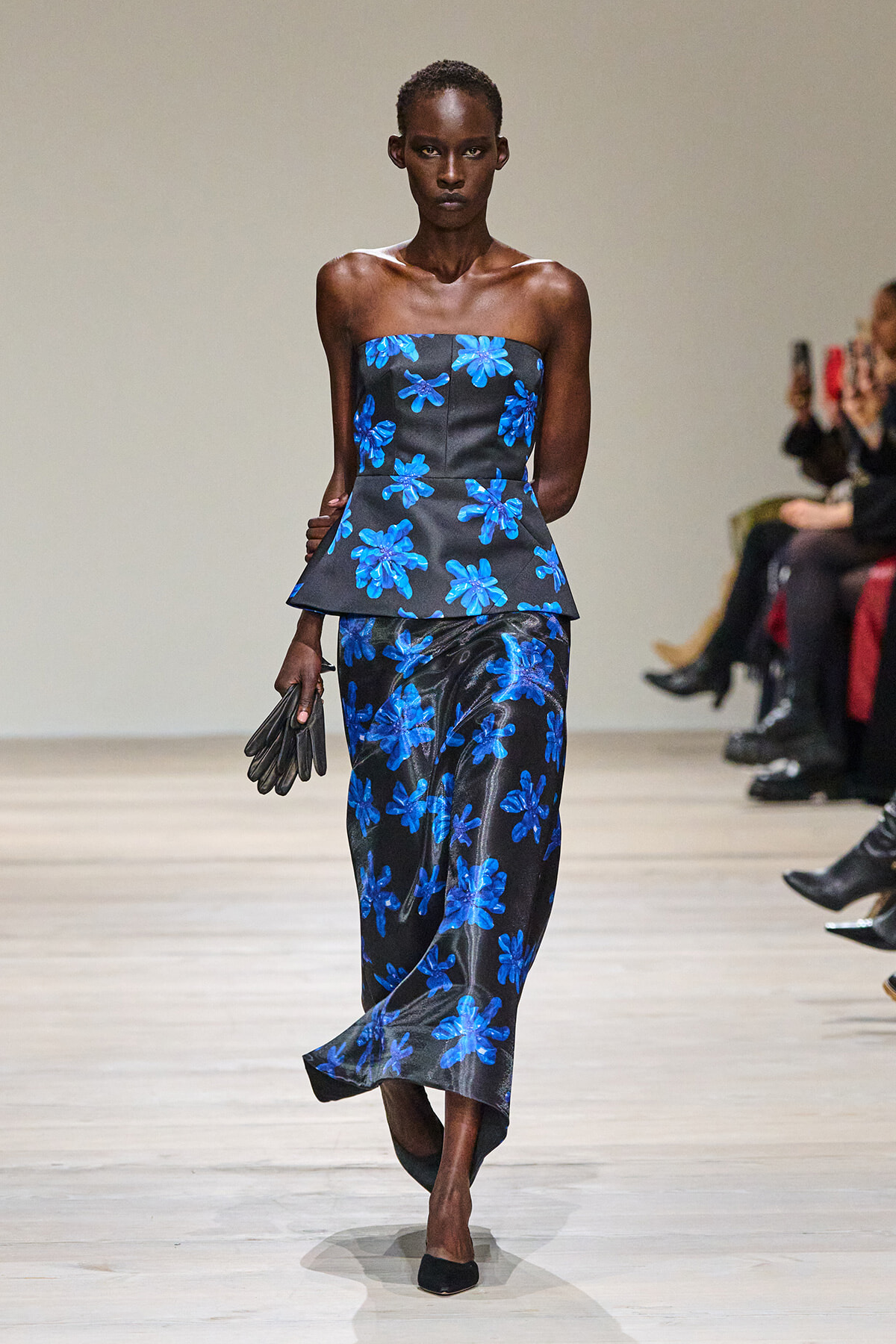 Model on the runway wearing a strapless black top with a blue floral print and matching skirt, holding black gloves.