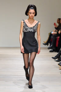 Model walking runway in a black cocktail dress with silver floral appliqués along the neckline, paired with sheer black tights and black heels, her dark hair styled in a sculpted updo.