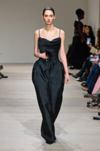 Model walking the runway in a black textured jumpsuit with thin spaghetti straps and a draped neckline, holding a small black accessory at the waist.