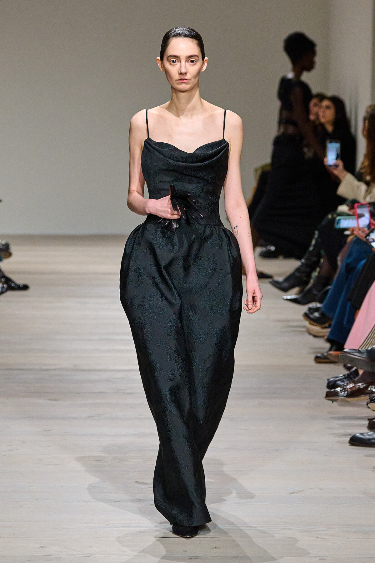 Model walking the runway in a black textured jumpsuit with thin spaghetti straps and a draped neckline, holding a small black accessory at the waist.