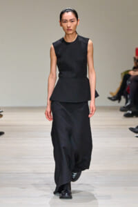 Female model on a runway wearing an all-black ensemble: sleeveless top with a peplum and a long, flowing skirt, paired with black boots.