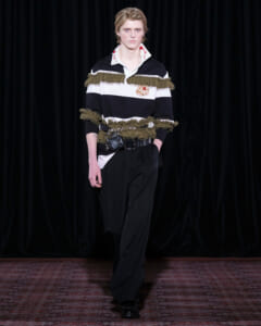 Male fashion model walking the runway in a black-and-white striped top with green fringe and crest embroidery, paired with black trousers and a belt bag.