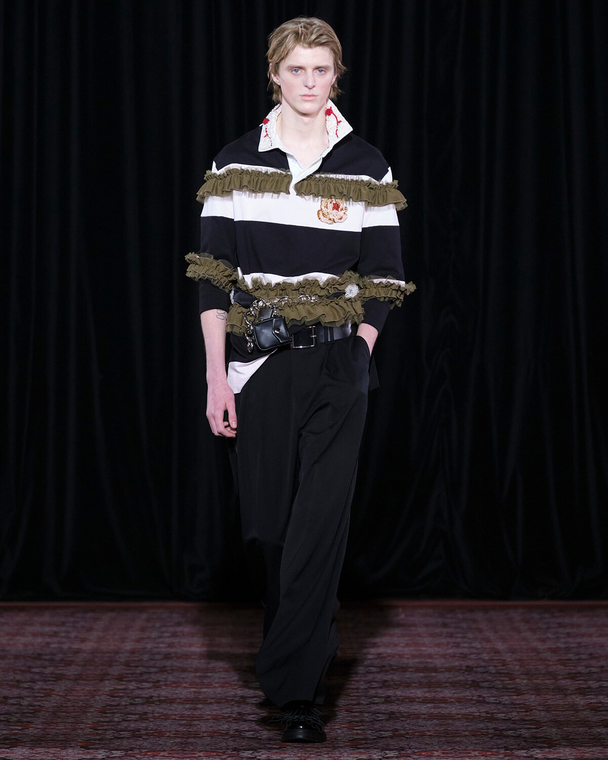 Male fashion model walking the runway in a black-and-white striped top with green fringe and crest embroidery, paired with black trousers and a belt bag.