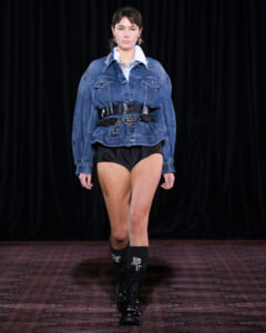 Model walking runway in oversized denim jacket cinched with a wide belt, black high-waisted shorts, and knee-high embellished boots against a dark backdrop.