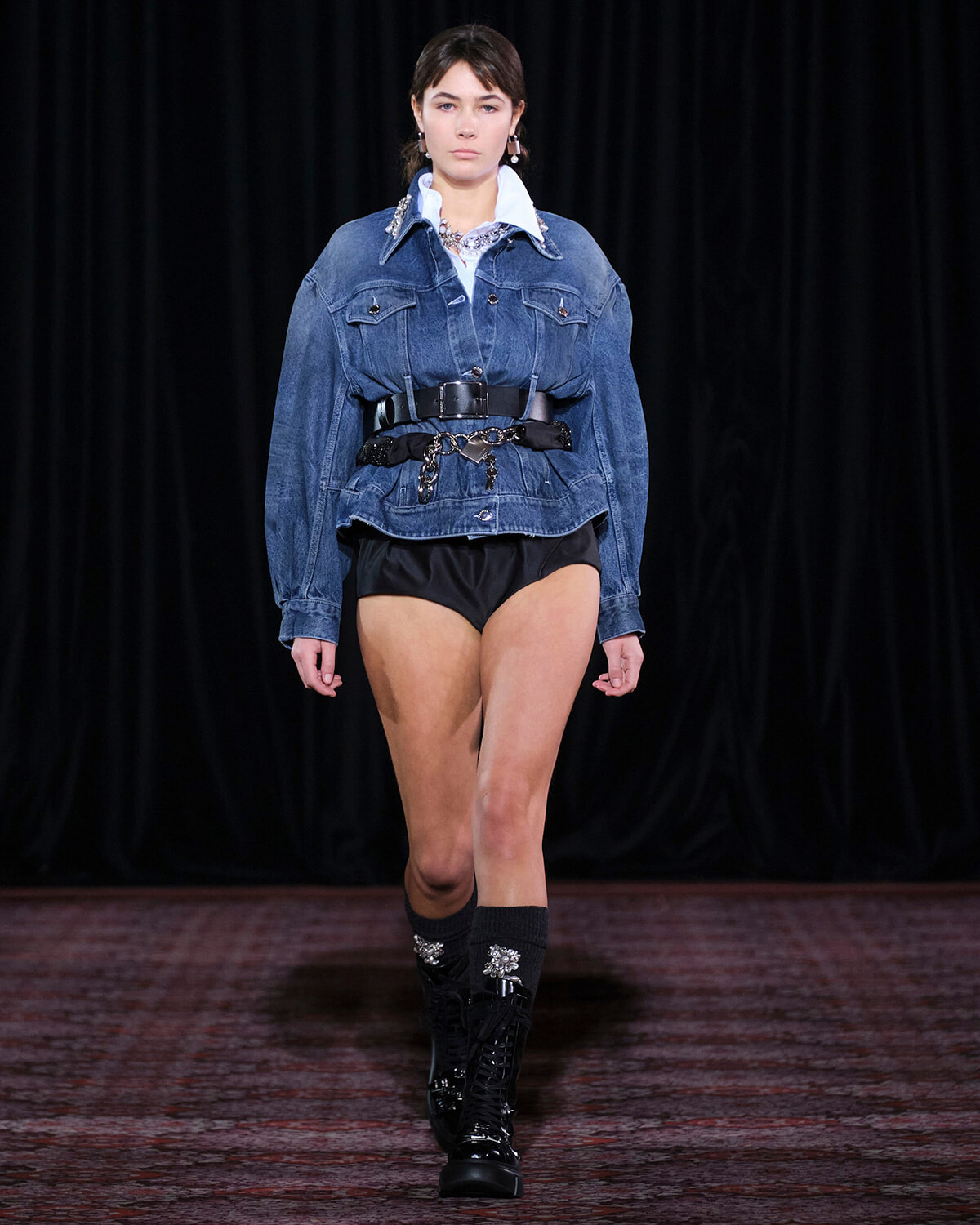 Model walking runway in oversized denim jacket cinched with a wide belt, black high-waisted shorts, and knee-high embellished boots against a dark backdrop.