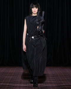Runway model wearing a black draped satin dress with long black gloves tucked under her arm, walking the catwalk against a dark backdrop.