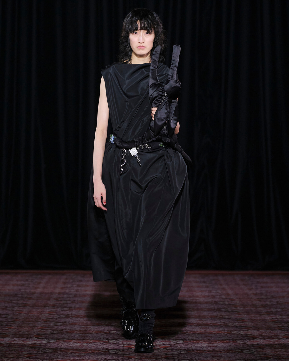Runway model wearing a black draped satin dress with long black gloves tucked under her arm, walking the catwalk against a dark backdrop.