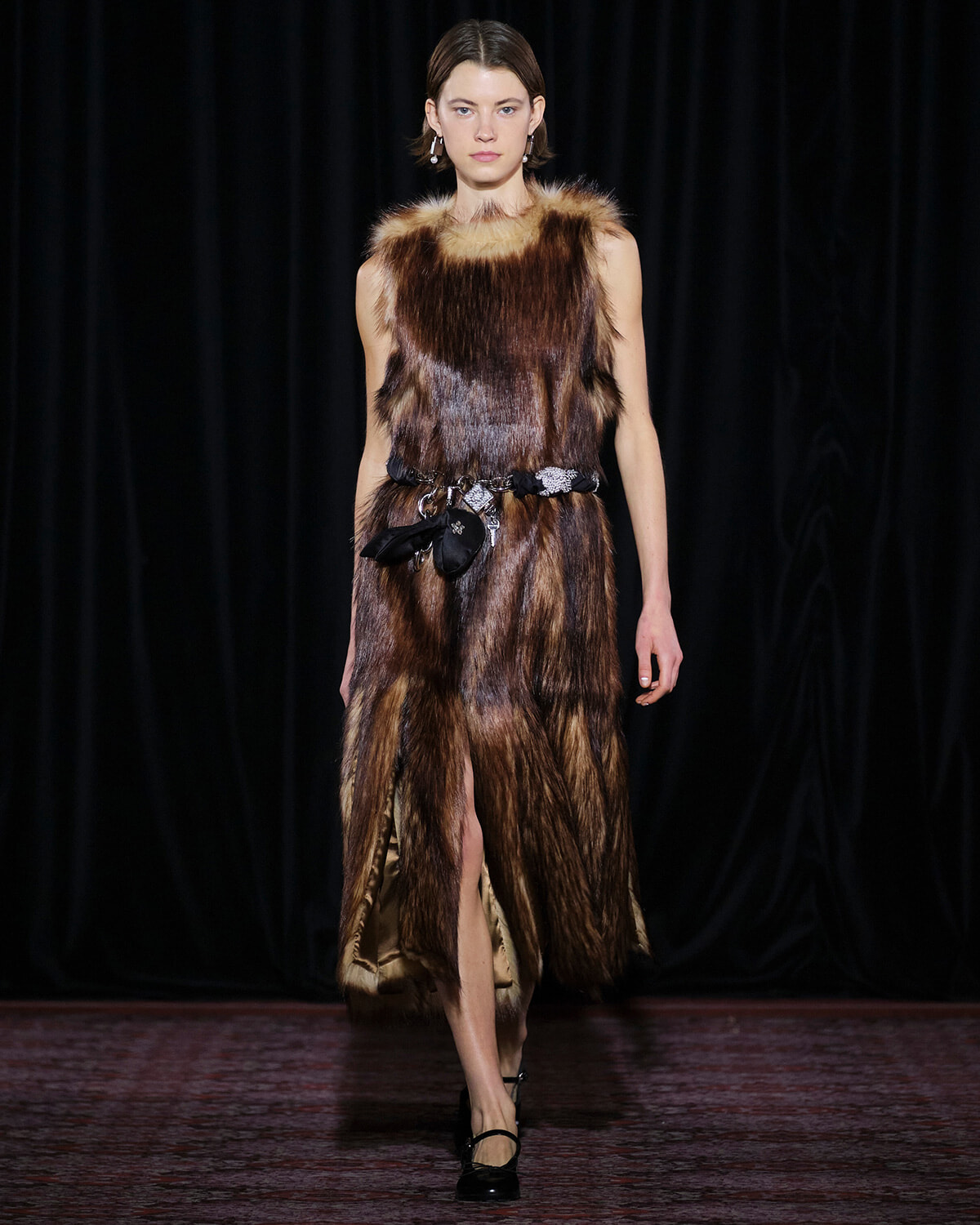 Model on a runway wearing a sleeveless brown fur dress, cinched with a decorative black belt and black heels.