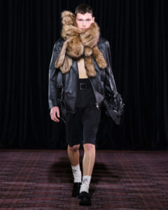 Model on a runway wearing a black leather jacket with an oversized fur scarf wrapped around the neck, paired with black shorts and loafers.