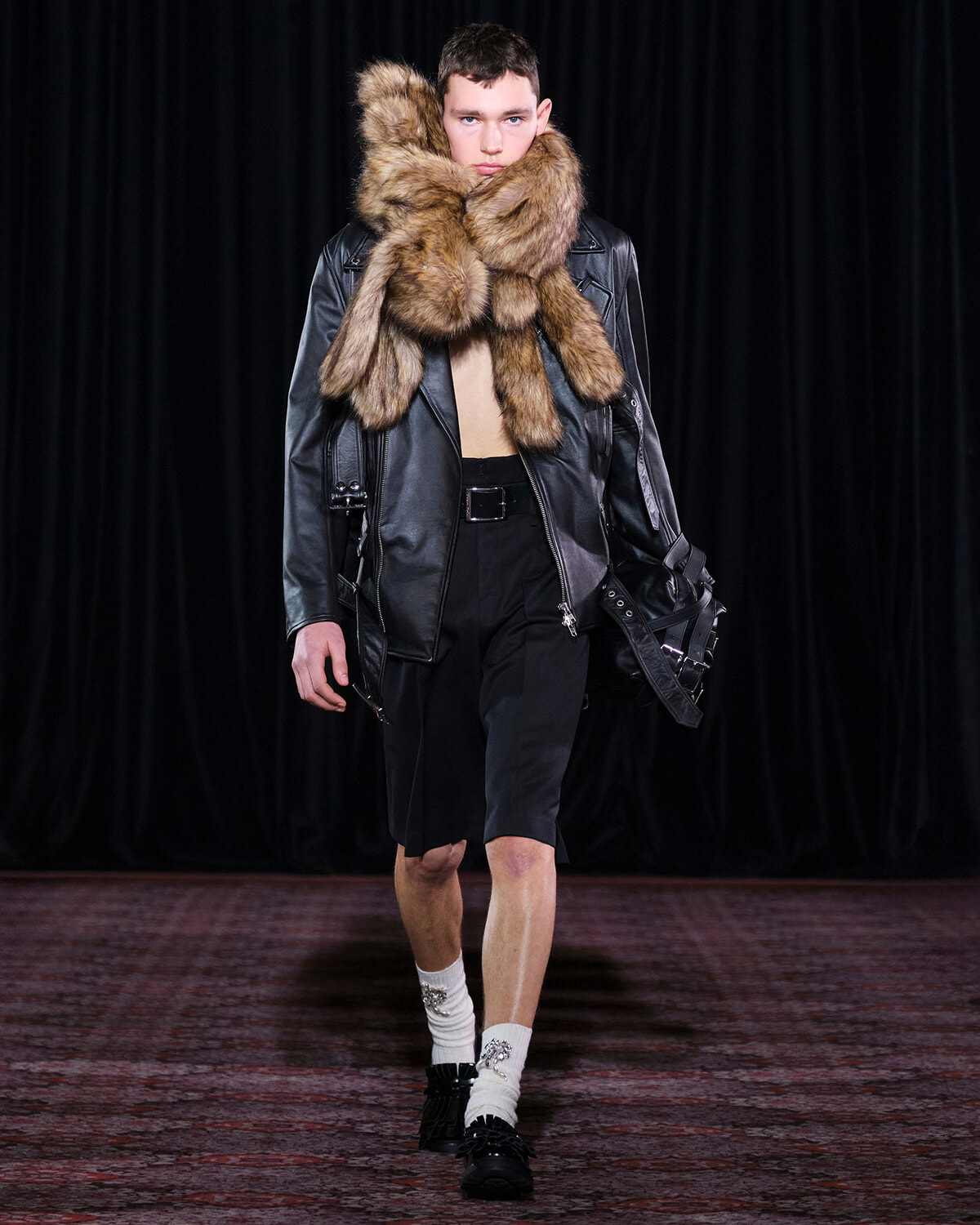 Model on a runway wearing a black leather jacket with an oversized fur scarf wrapped around the neck, paired with black shorts and loafers.