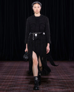 Model walking a glamourous black textured dress with a chain belt and headband on a runway, carrying a small black handbag.