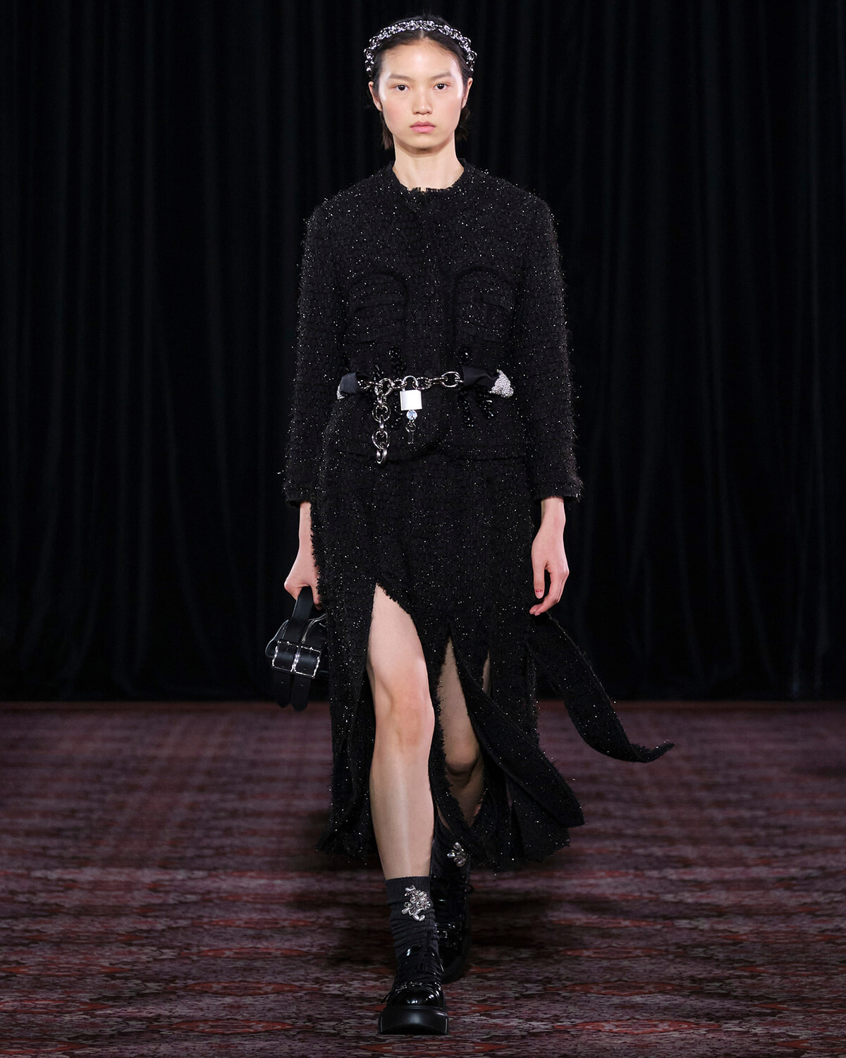 Model walking a glamourous black textured dress with a chain belt and headband on a runway, carrying a small black handbag.