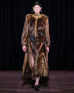 Model walks a runway in a long brown fur coat with a belt bag and black shoes, against a dark backdrop.