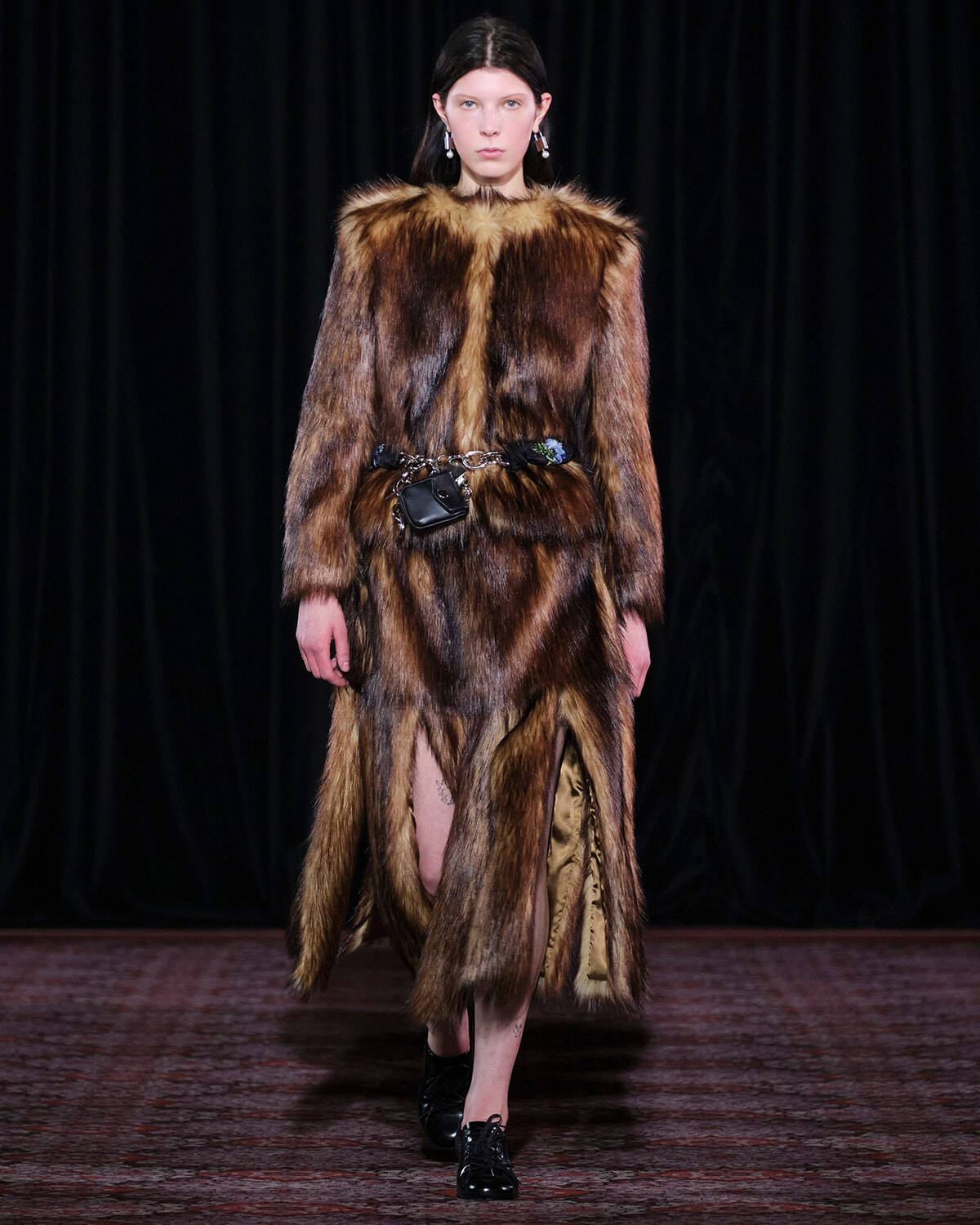 Model walks a runway in a long brown fur coat with a belt bag and black shoes, against a dark backdrop.