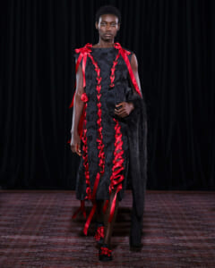 Model on a runway wearing a black sleeveless dress with red satin ribbons braided down the front, holding a black clutch and fur stole.