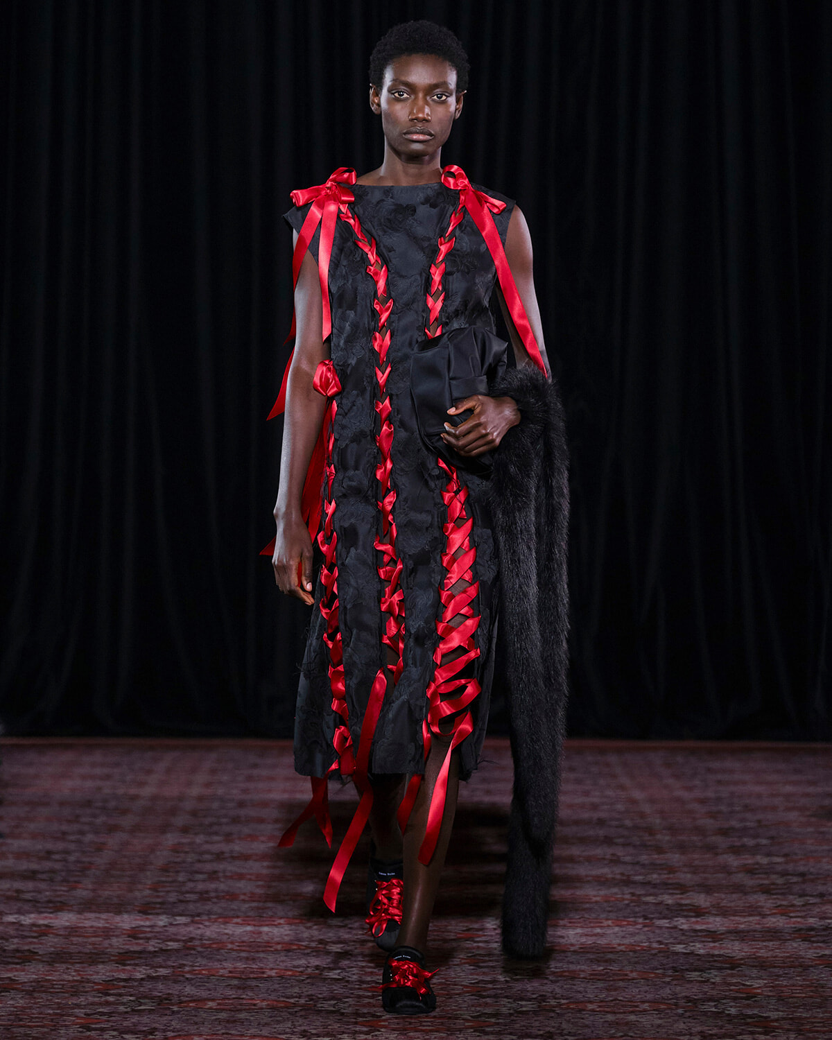 Model on a runway wearing a black sleeveless dress with red satin ribbons braided down the front, holding a black clutch and fur stole.