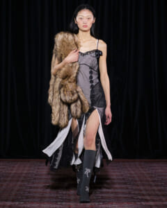 Runway model walking in a gray satin dress with black lace, holding a large fur stole over her shoulder and a high slit showing her legs.