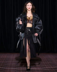 Model walking a runway in a black leather oversized jacket, fur crop top, and a high-slit black skirt with a chain belt, carrying a black bag.