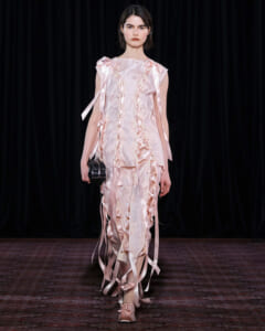Female model walks the runway in a pale pink, ruffled gown with satin ribbons and a small black clutch.